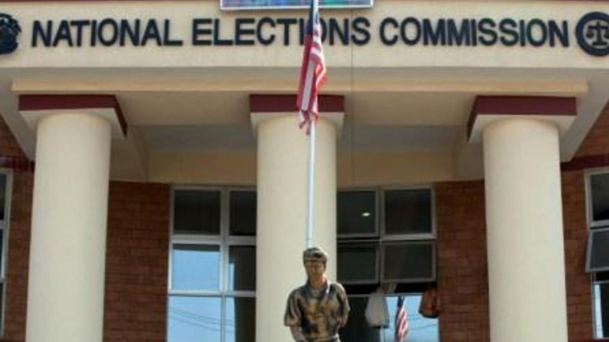 A peacekeeper standing in front of a flag pole flying the Liberian flag and in front of the Liberian National Elections Commission.
