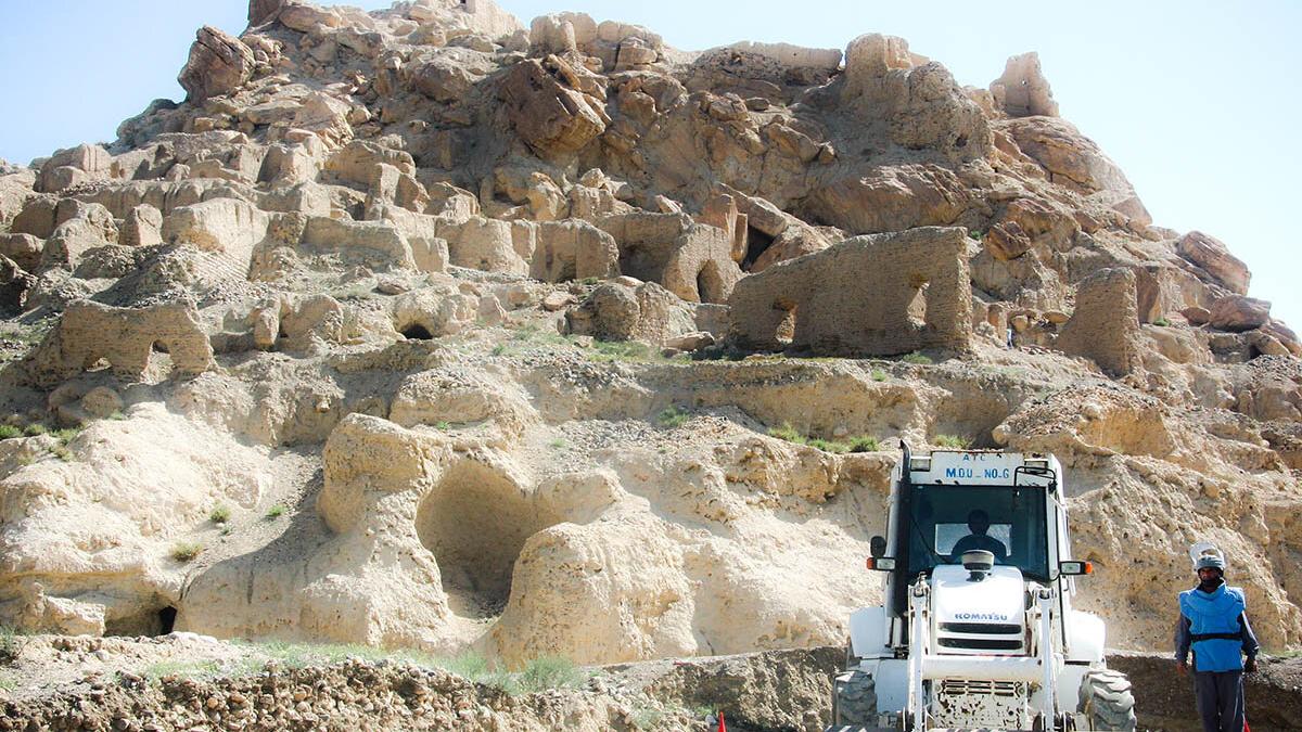 A rocky hillside with ancient-looking stone ruins carved into the cliff face, while a white construction vehicle and a worker stand at the base