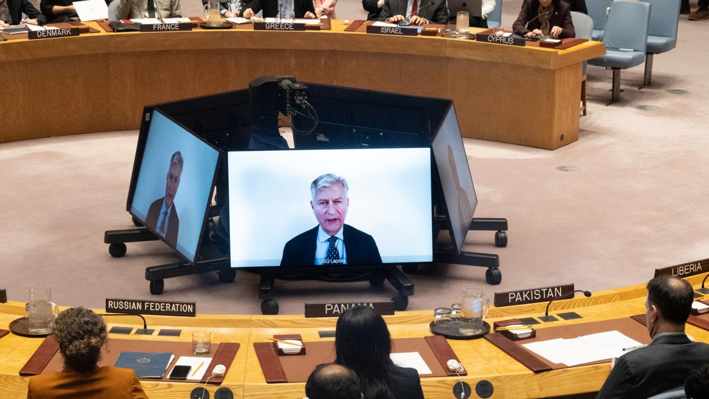 A Security Council meeting room with delegates seated around a circular table, nameplates visible for several countries. At the center, multiple large monitors display a remote briefing speaker. Attendees are focused on the screens, and documents, microphones, and translation earpieces are arranged on the desks.