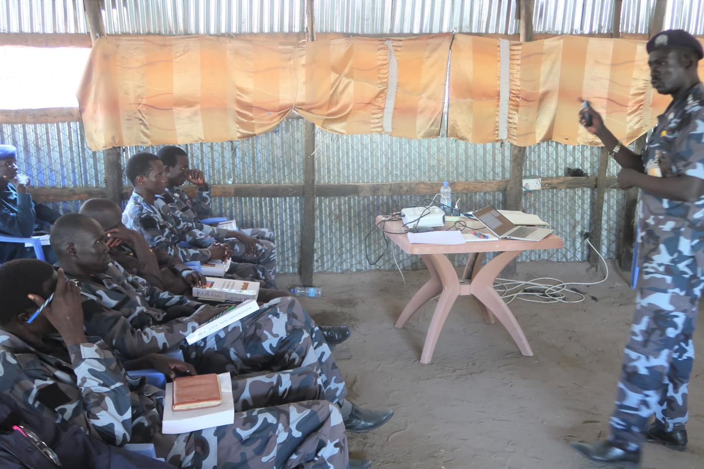 A group of uniformed police officers sitting watching a presentation being given by another uniformed person.