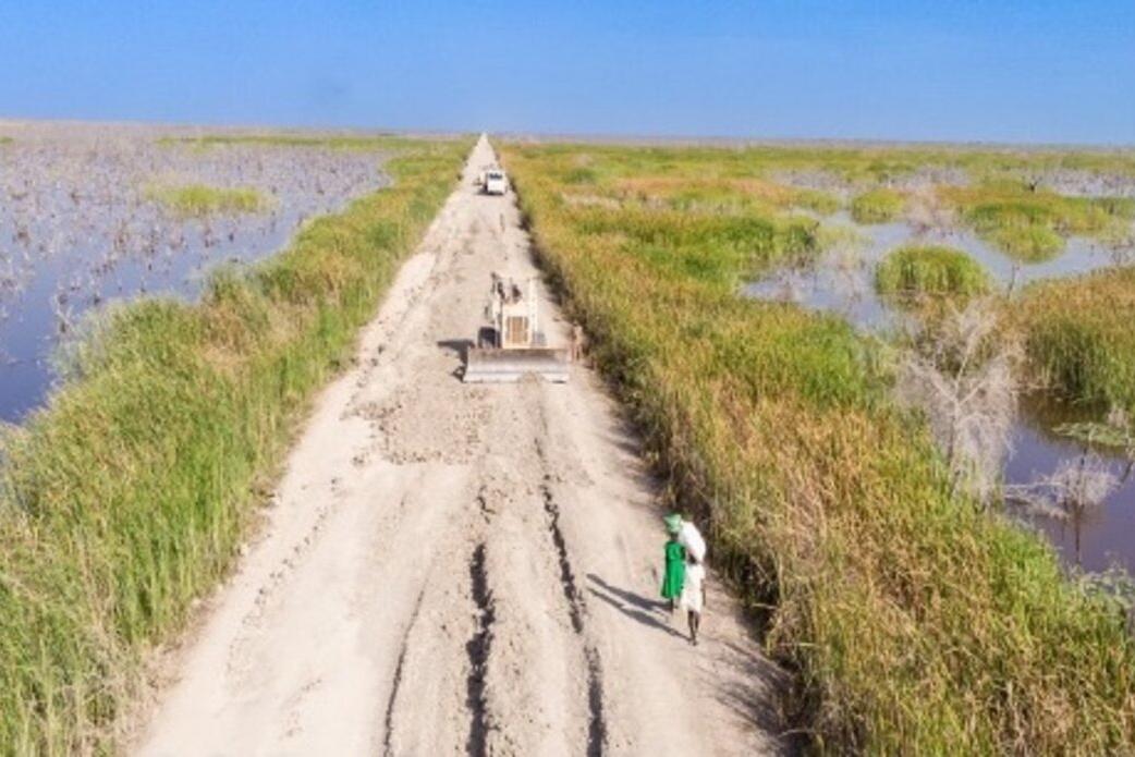 An aerial view of a dirt road with grass and bodies of water on either side. On the road, there are some people walking. 