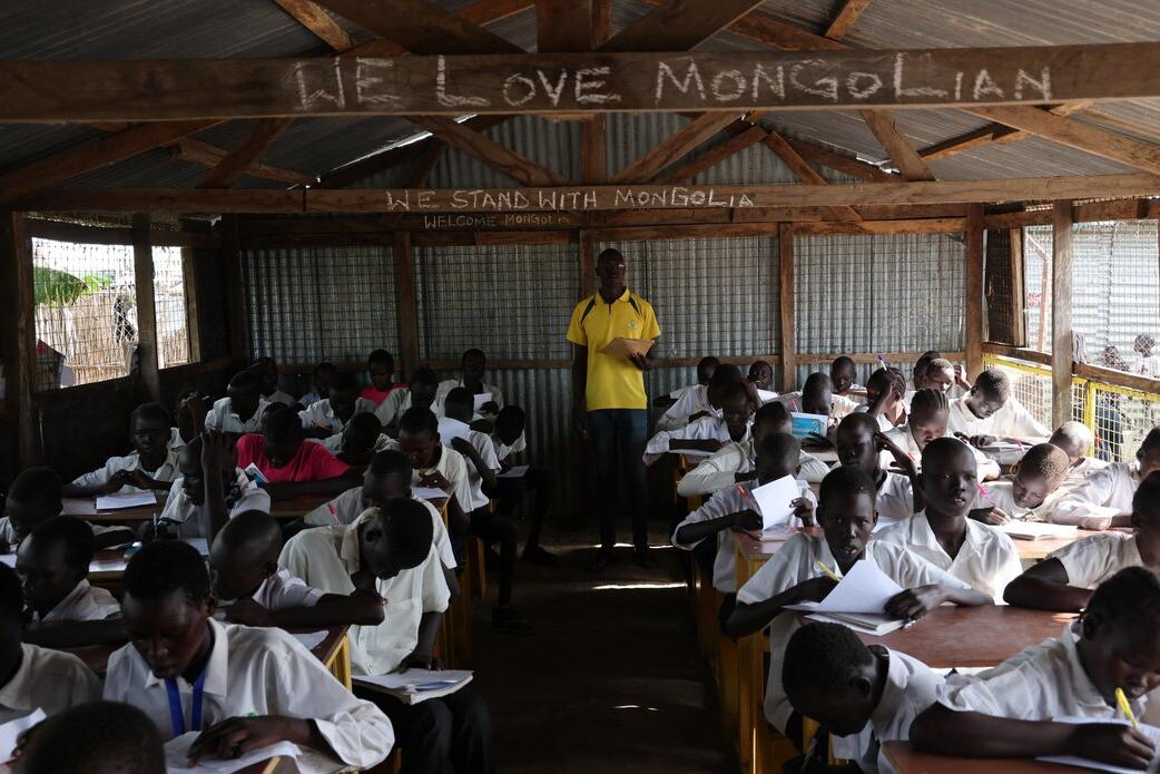 A classroom with students in white uniforms seated at wooden desks, writing in notebooks, while an instructor stands at the front under a corrugated metal roof with chalk writing above.