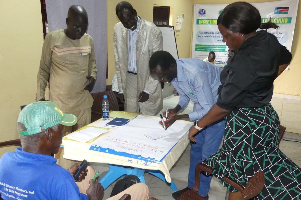 Group of people gathered around a table indoors, reviewing and writing on large sheets of paper during a workshop or planning session.