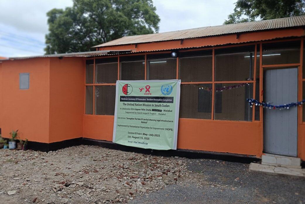 An orange building with a banner displaying information about a UN mission and HIV awareness program, with a door and windows visible and trees in the background.