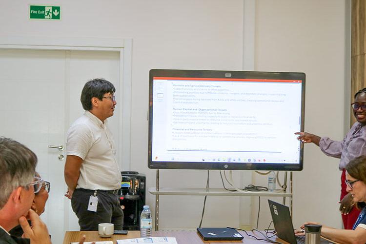 A group of people in a meeting room are gathered around a large digital screen displaying text. One person stands and points at the screen, while another stands nearby. Two others are seated at the table with laptops and documents in front of them. The room has a white wall with a green exit sign and a wooden panel accent.