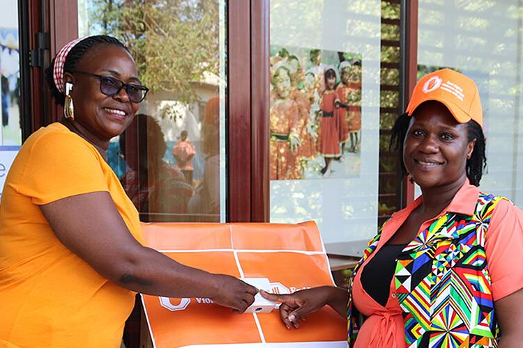 Two women both wearing orange coloured clothing and smiling each putting a finger on a button between them