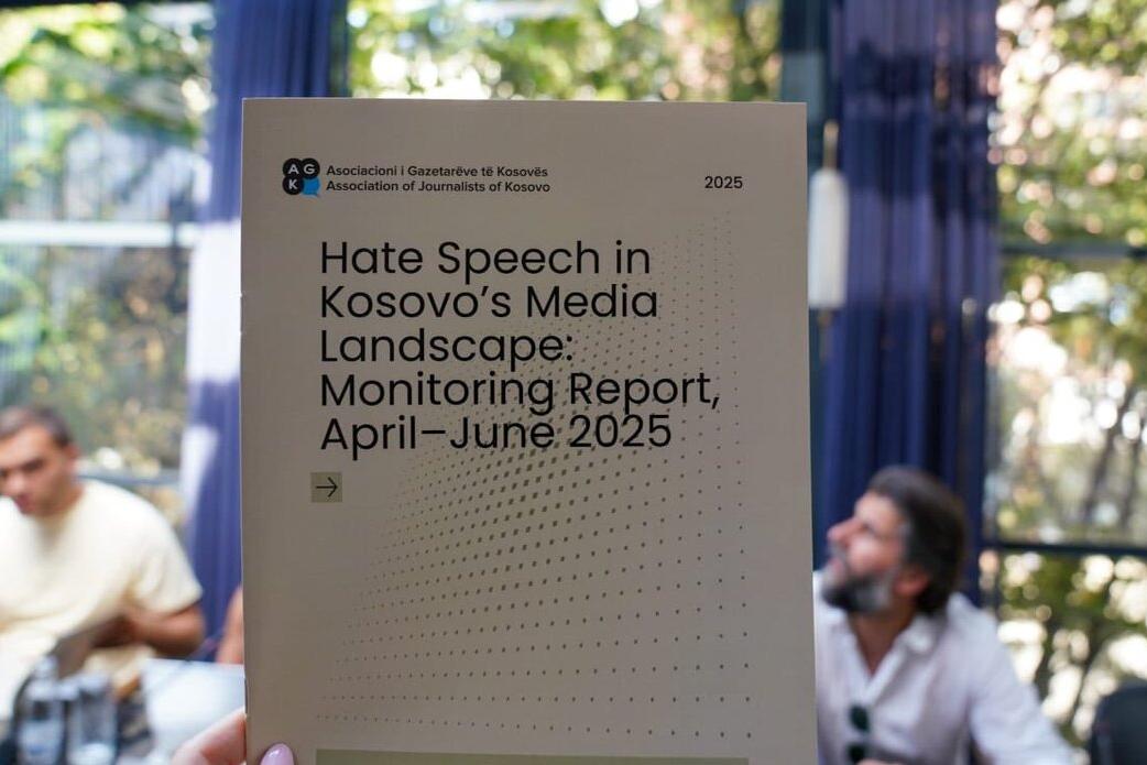 A close-up of a hand holding a report titled “Hate Speech in Kosovo’s Media Landscape: Monitoring Report, April–June 2025” published by the Association of Journalists of Kosovo. The background shows a meeting setting with people seated at a table and large windows with greenery outside.