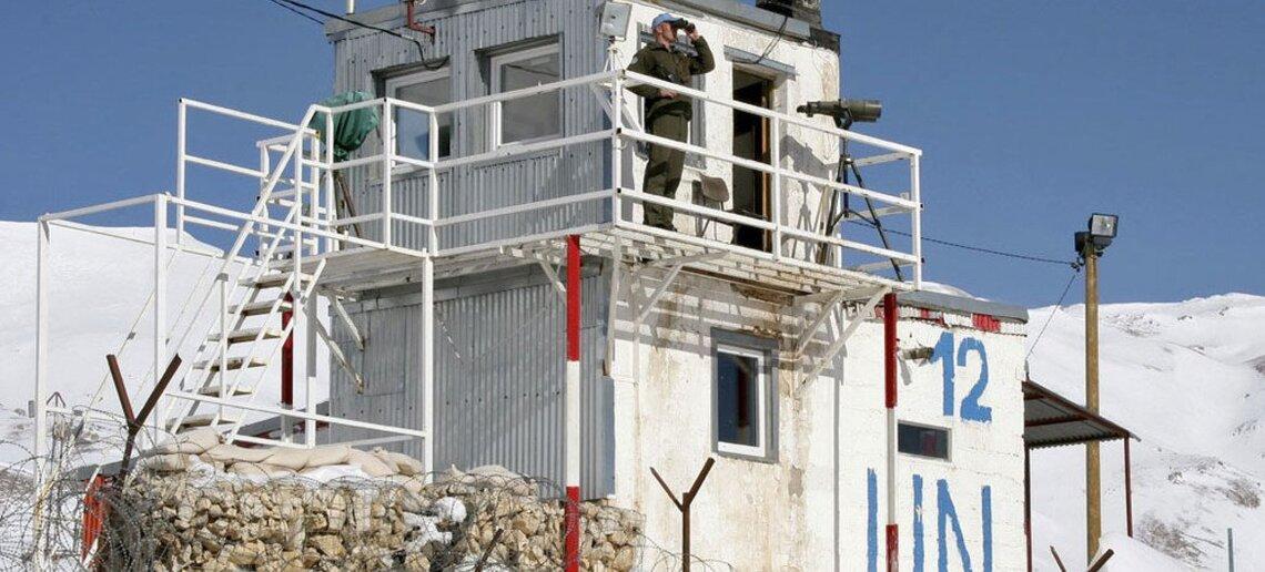 A peacekeeper stands on top of a UNDOF observatoin tower, looking through binoculars into the distance.