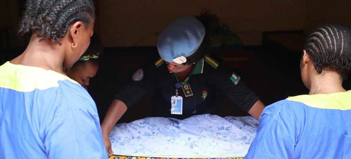 Four individuals gathered around a table with a blue and white patterned cloth, one in a uniform with a blue beret and ID badge instructing three others dressed in blue garments with yellow accents, in a dimly lit indoor setting
