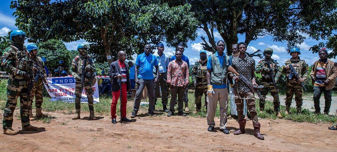 Men in civilian clothing and in peacekeeping uniforms stand outside, some carrying guns. 