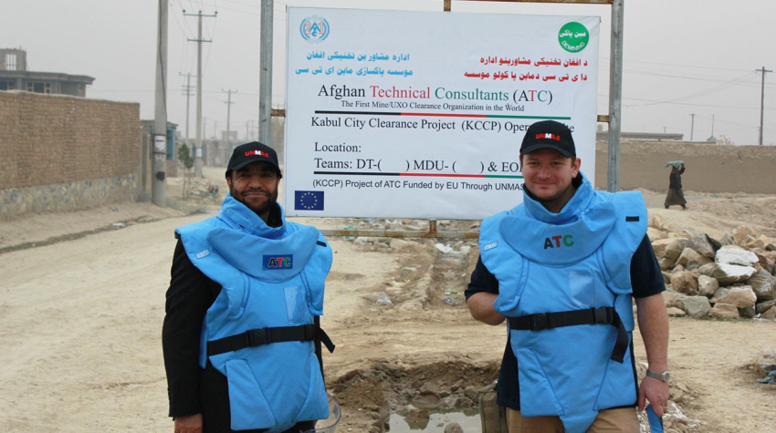 Two people in ATC protective gear stand in front of a Kabul City Clearance Project sign, with buildings and a dirt road in the background.