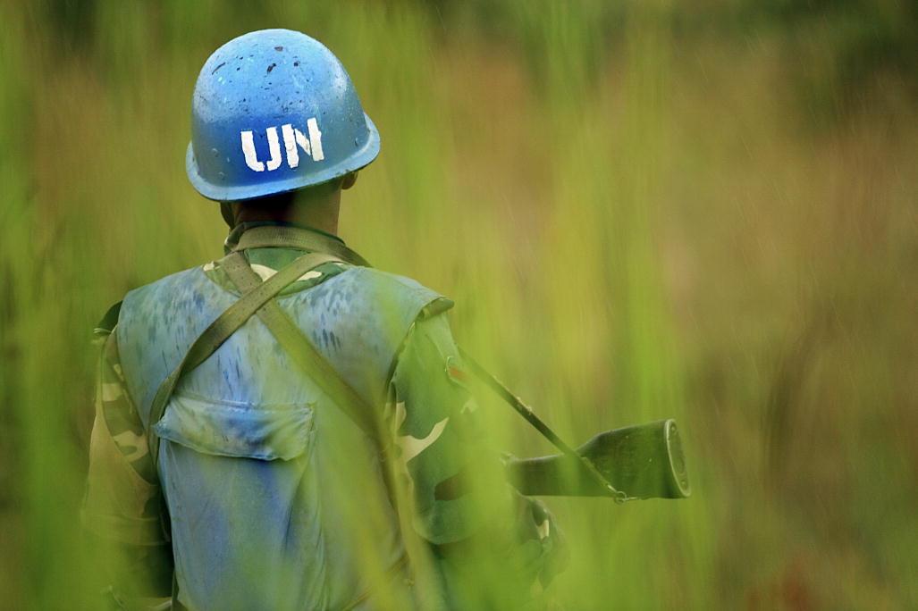 A UN peacekeeper in a blue helmet stands with his back to the camera in tall grass